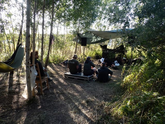 Figures sitting in dappled light beneath trees, tarpaulin and speaker stand behind them.