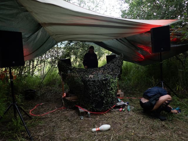 Night set under a tarpaulin canopy, speakers on stands and red light through the trees.